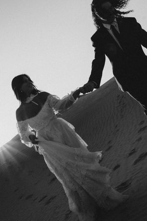 Black and white photo of bride and groom holding hands on sandy desert dunes, sun flare behind flowing lace wedding dress.