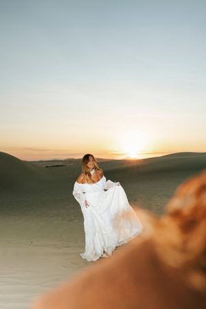 Bride in white lace off-shoulder gown twirling on desert sand dunes at sunset.