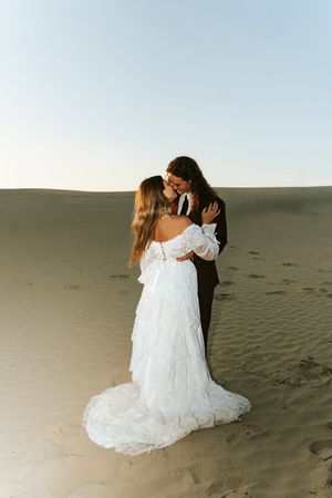 Bride and groom embracing on sandy desert dunes at golden hour sunset.