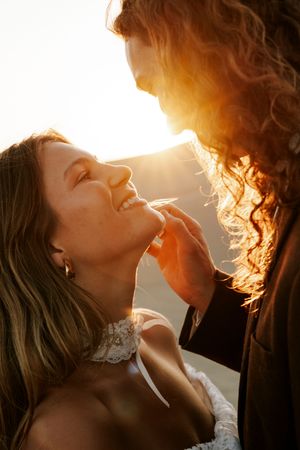 Bride and groom share a romantic golden sunset moment, with groom tenderly touching brides chin in warm backlit glow.