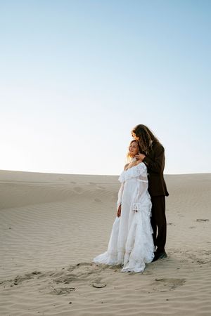 Bride in white lace off-shoulder gown embraced by groom in black suit on serene desert sand dunes at golden hour.
