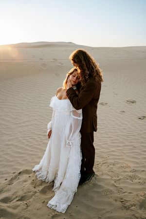 Bride in white lace dress and groom in brown suit embrace tenderly on sandy desert dunes at golden sunset.