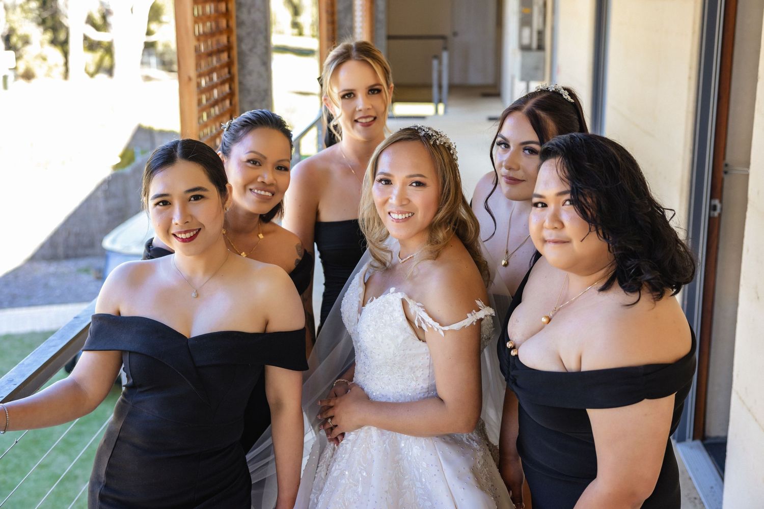 Bride in white gown poses with five bridesmaids in black off-shoulder dresses outdoors at a wedding venue.
