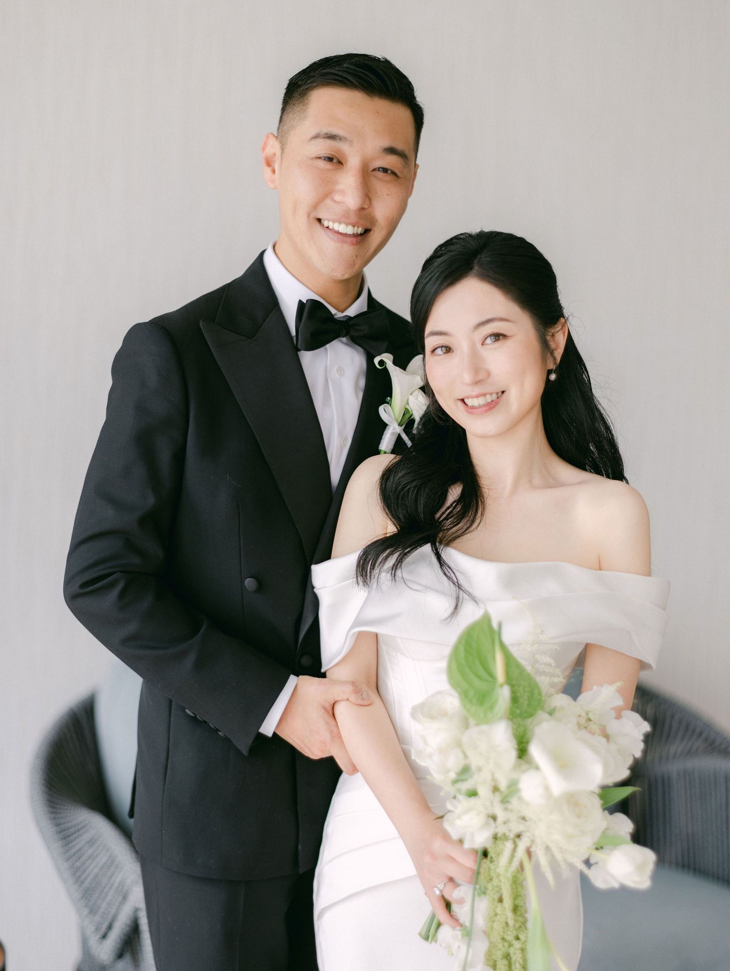 Smiling couple in wedding attire, groom in black tuxedo and bride in off-shoulder white gown holding white bouquet.