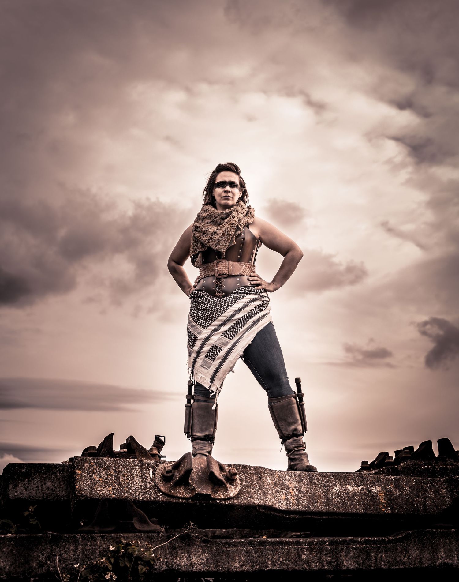 A warrior woman stands boldly atop ruins under dramatic cloudy skies, wearing tribal costume with armor leg guards.