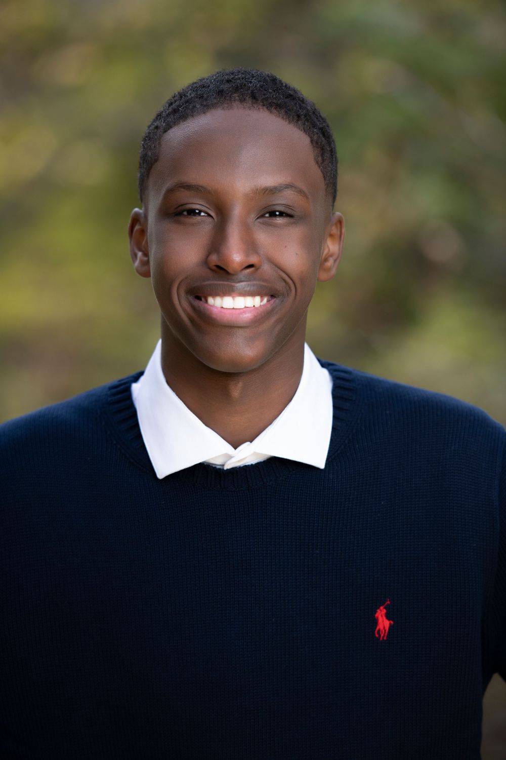 Smiling young man in navy Ralph Lauren sweater over white collar shirt, outdoor portrait with green background.