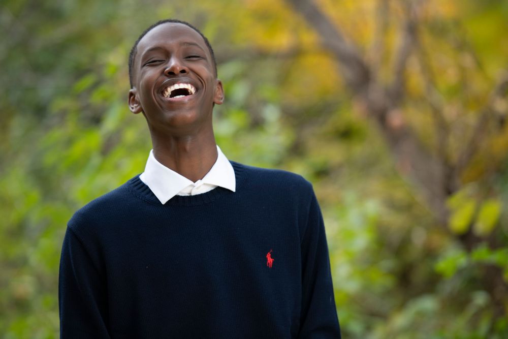 Young Black man in navy Ralph Lauren sweater laughing joyfully outdoors with autumn foliage background.
