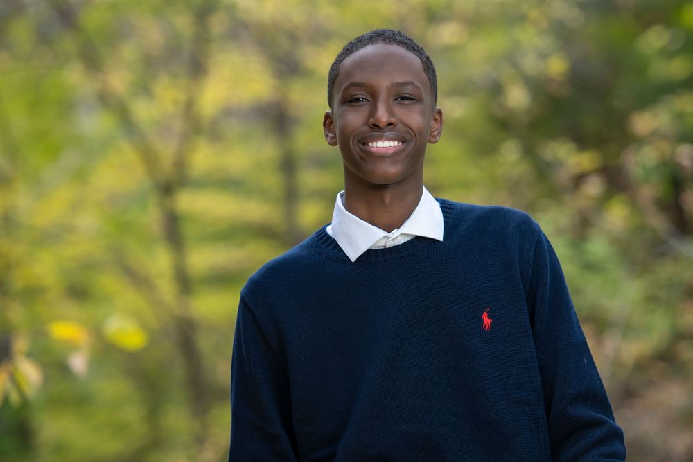 Young man smiling outdoors in navy Ralph Lauren sweater with autumn foliage background.