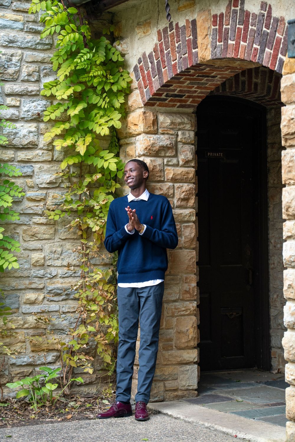 Young man in navy sweater and gray trousers stands smiling by a stone archway with ivy, clapping hands.
