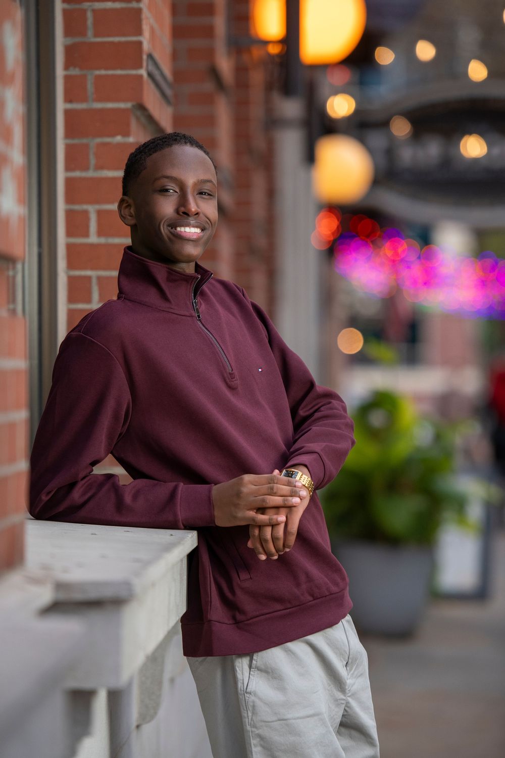 Smiling Black man in maroon hoodie leaning on railing at urban outdoor venue with bokeh lights.