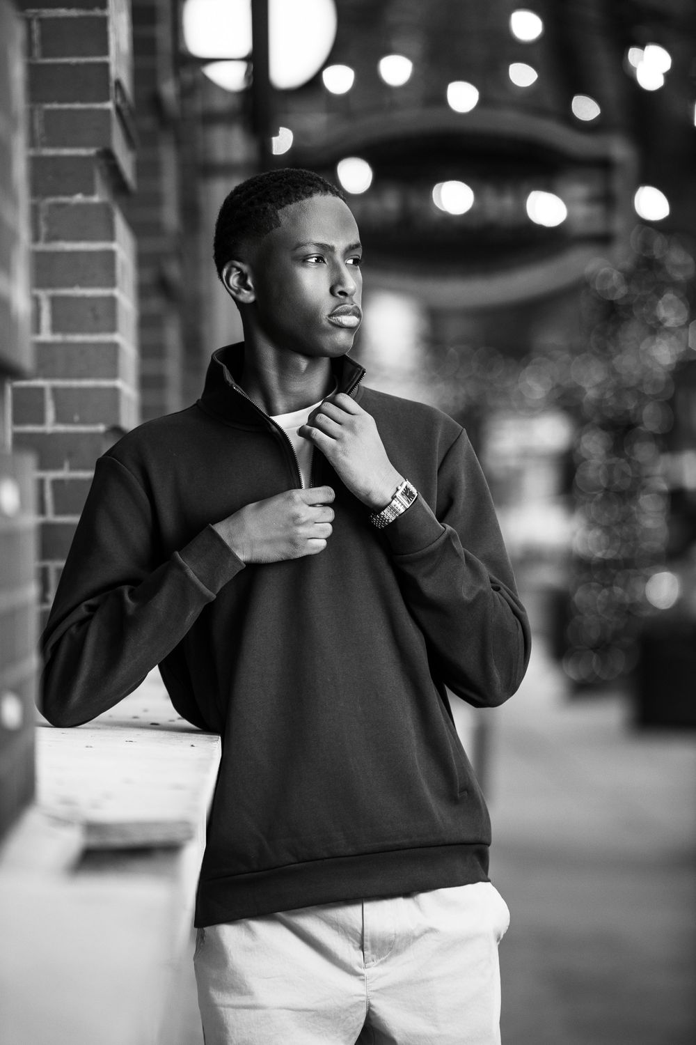 Black and white portrait of a young man in a quarter-zip sweatshirt leaning against a brick wall outdoors.