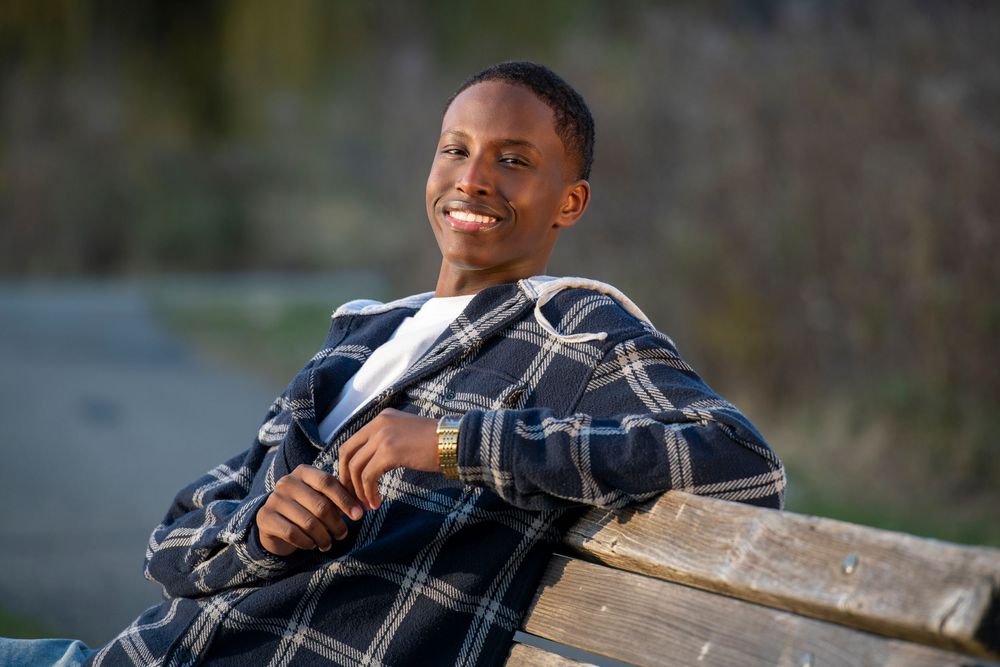 Smiling young man in plaid blazer sitting relaxed on a wooden bench outdoors in natural light.