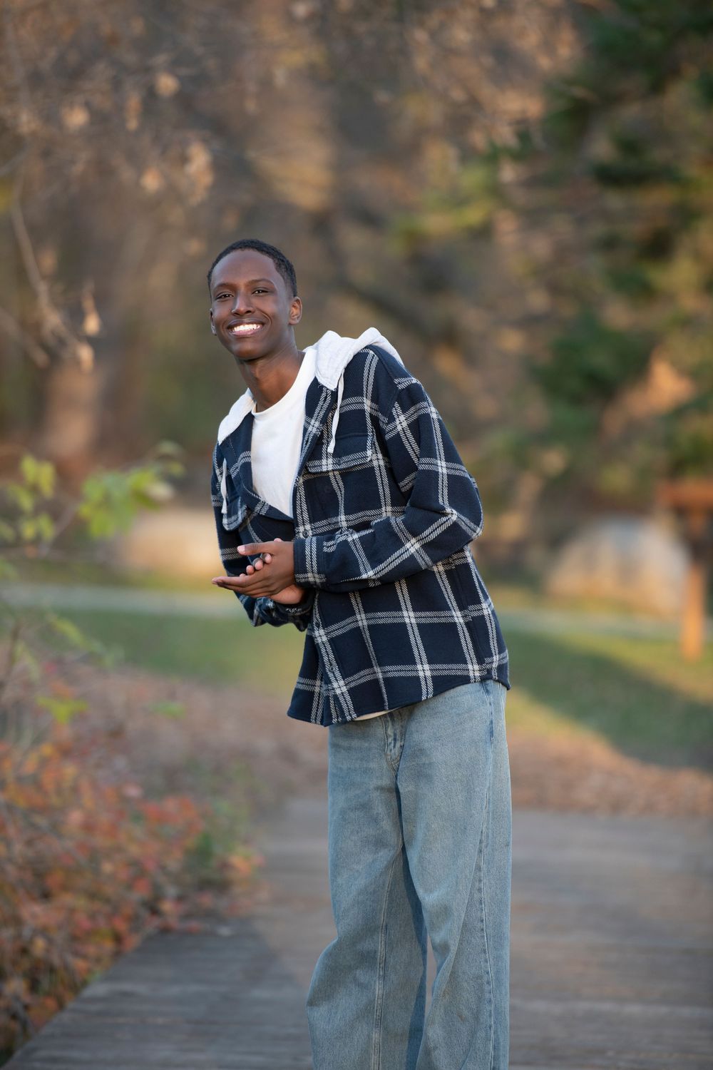 Smiling Black man in plaid shirt and jeans holding camera on outdoor park path with autumn trees.
