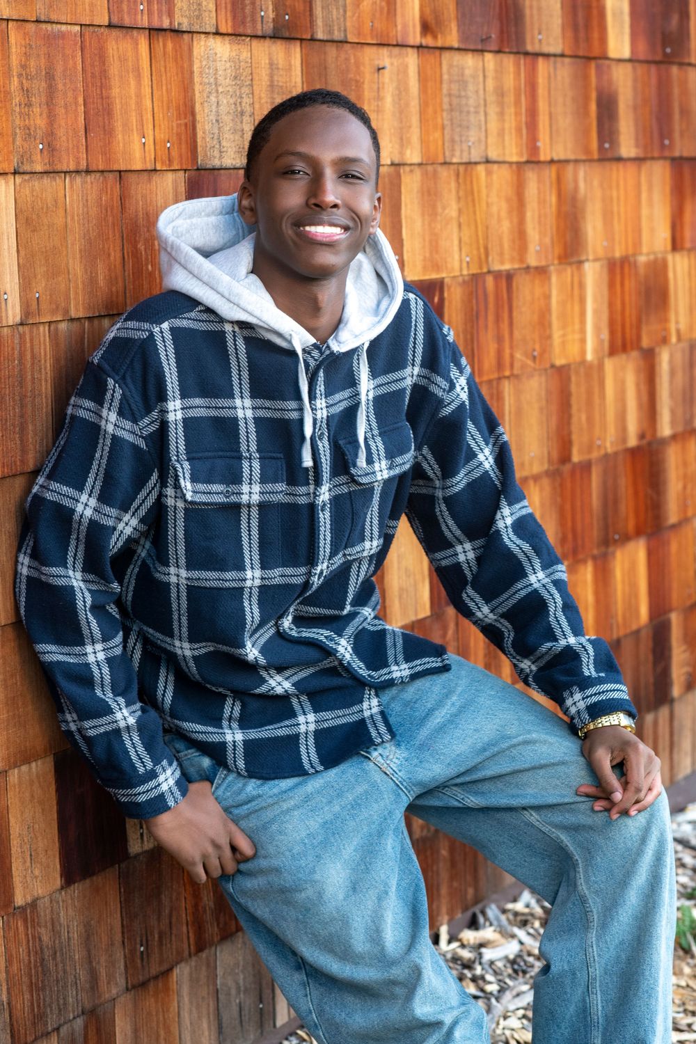 Young Black man smiling, wearing plaid flannel hoodie and jeans, leaning against rustic wooden wall outdoors.