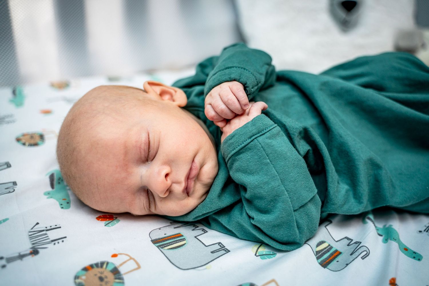 Peaceful newborn baby sleeping in a green outfit on a colorful animal-print blanket in a crib.