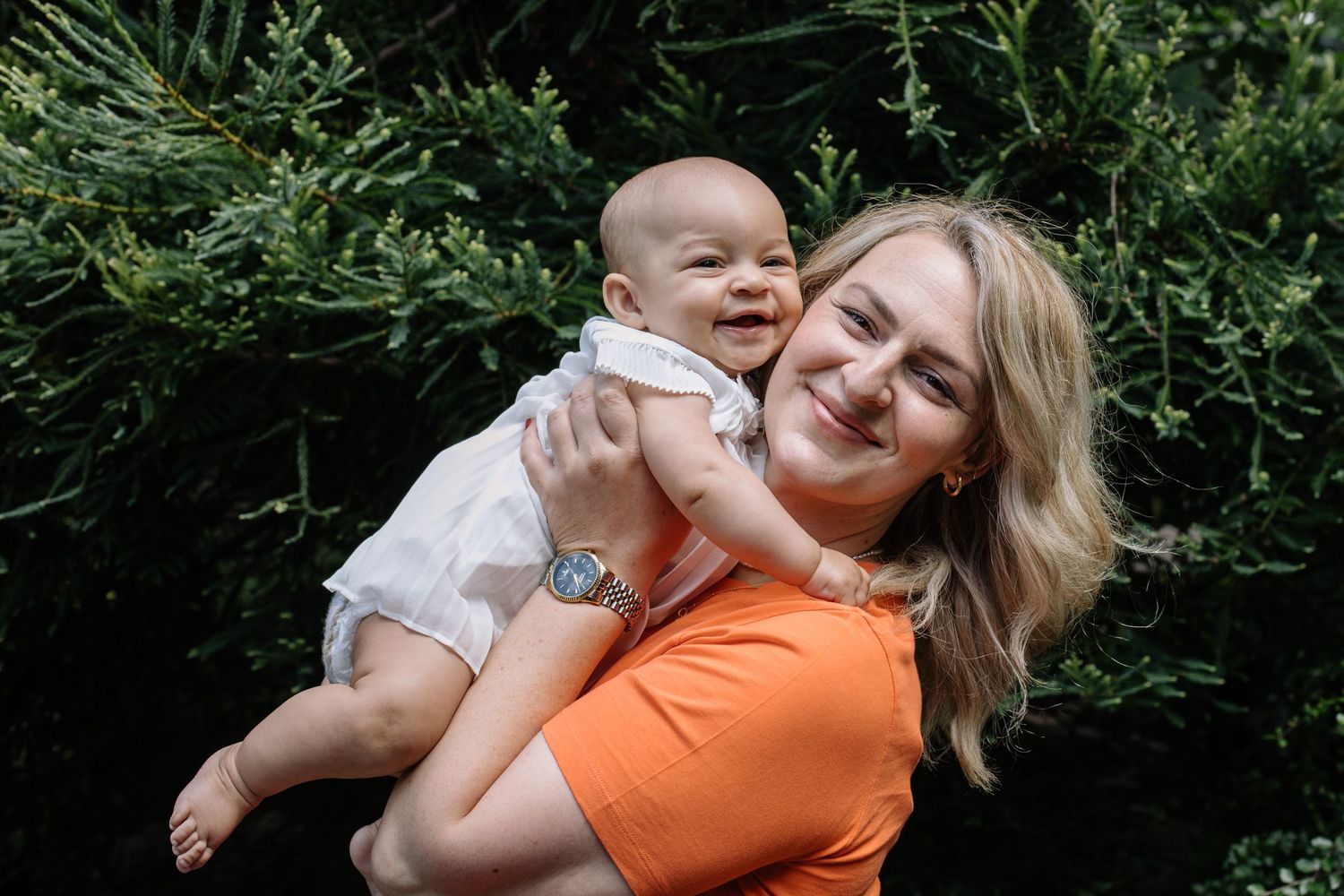 A smiling woman in an orange top holds a happy baby outdoors with lush green bushes in the background.