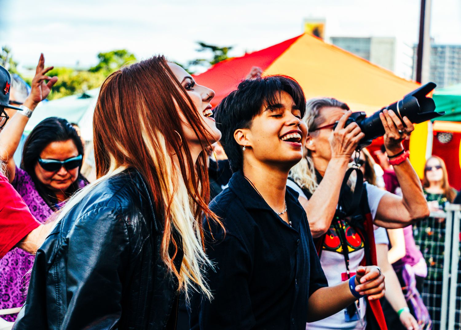 A diverse crowd of festival attendees enjoying an outdoor event with colorful umbrellas and drinks in hand.