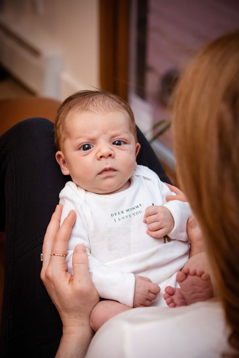 Baby in white onesie being held during indoor portrait session with natural lighting.