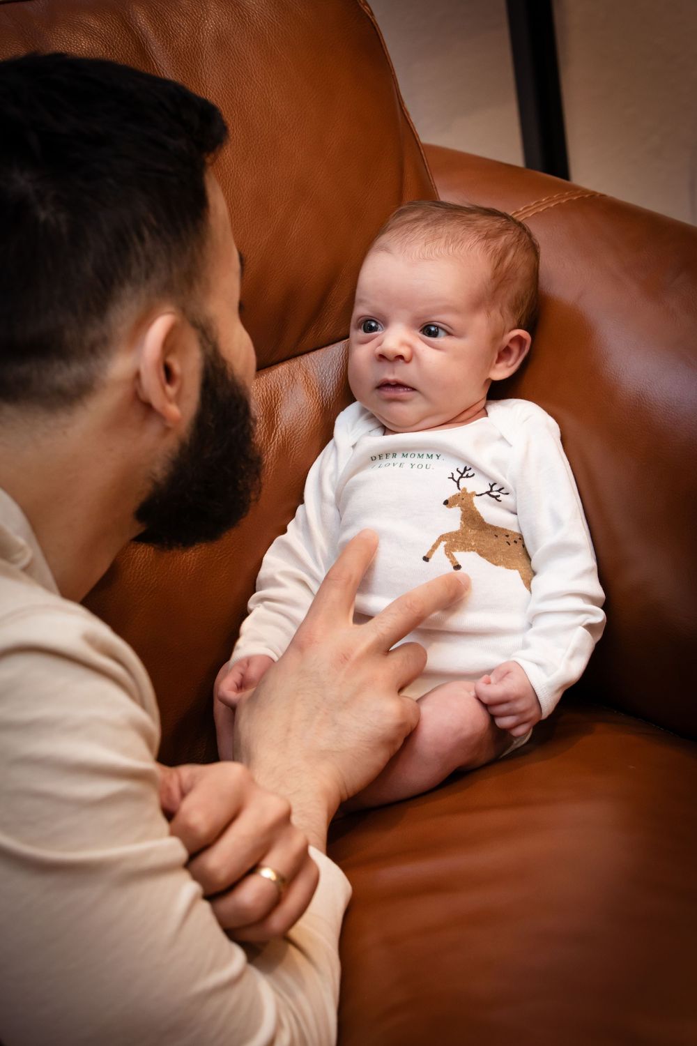 Baby in white onesie looking alert during casual indoor photo session on leather couch.