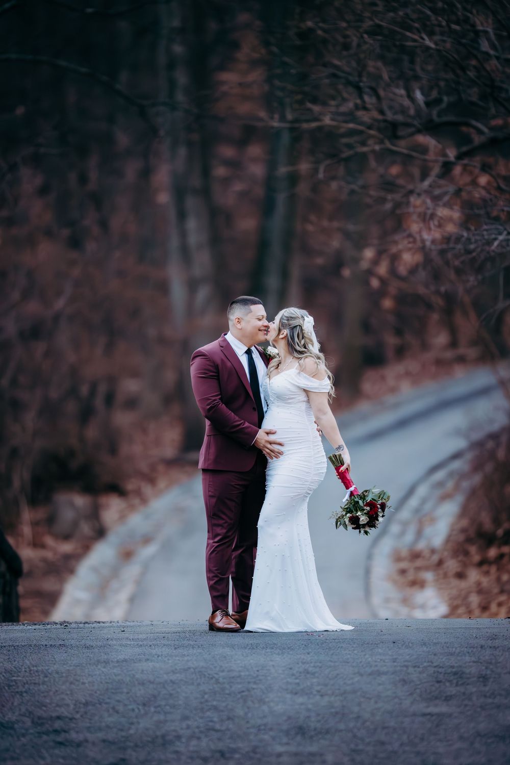 A romantic wedding photo of a couple on a tree-lined road with autumn colors and burgundy accents.