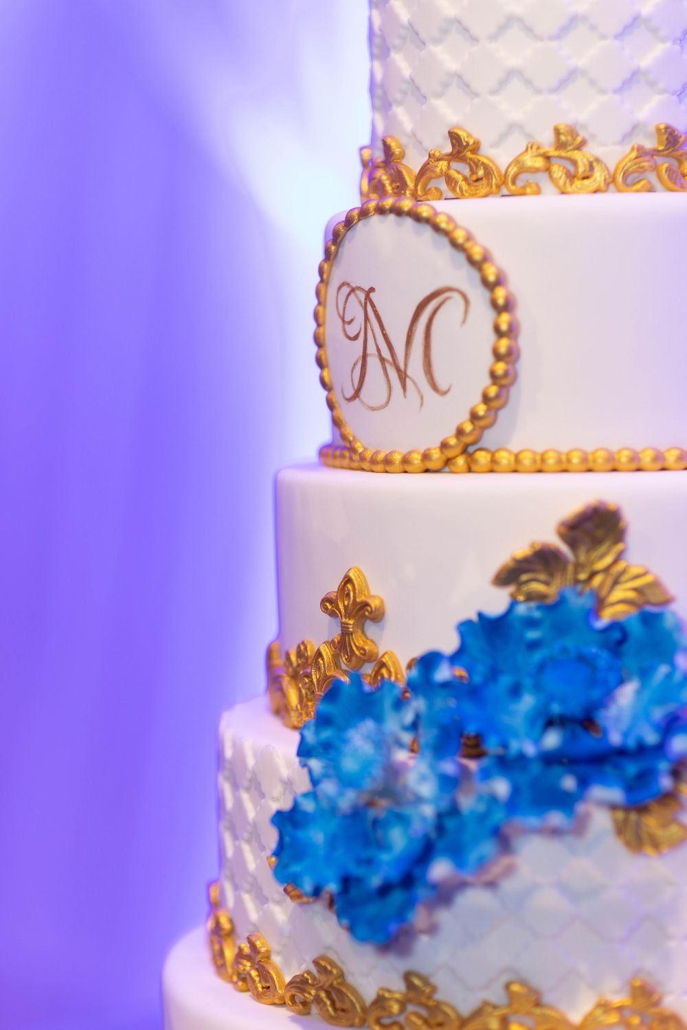 Elegant white tiered cake with blue flowers and gold accents photographed against purple lighting.