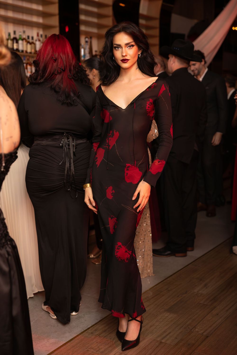 A figure in a black dress with red floral accents poses dramatically at a dimly lit social gathering.