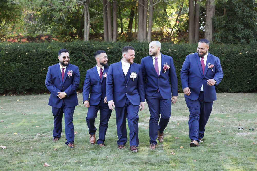 Groomsmen in matching navy suits walk together on a grassy lawn.