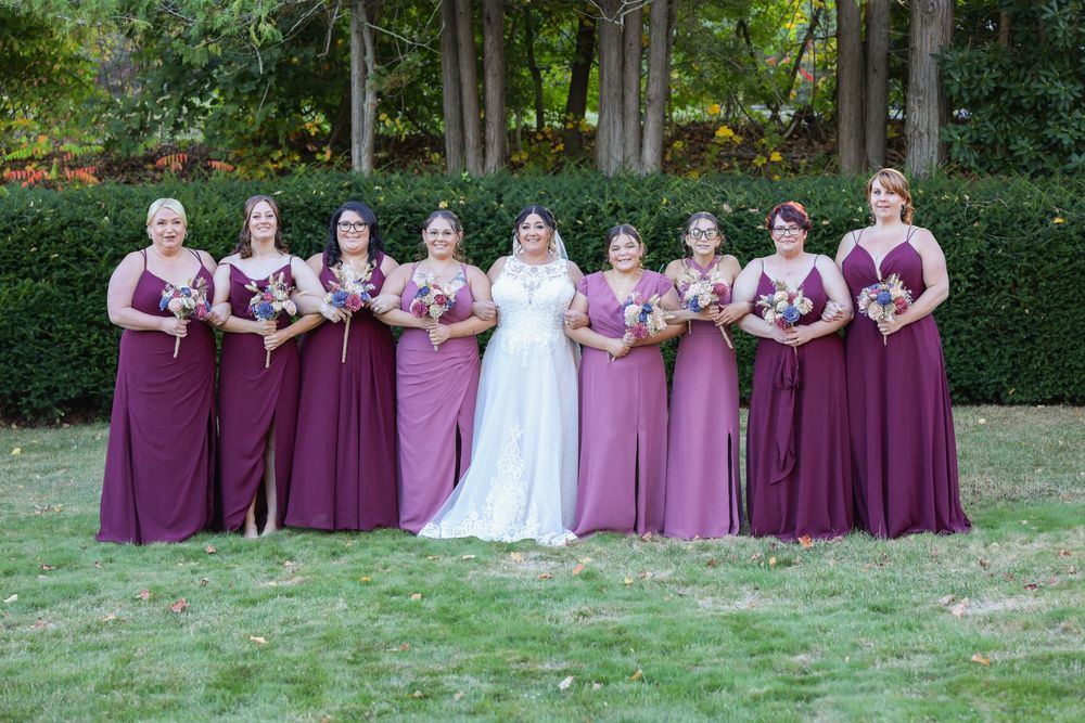 Wedding party in purple bridesmaid dresses poses on a lawn with trees in background.