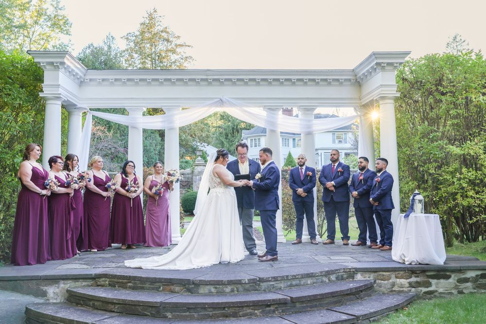 Wedding party poses for formal photos in front of white gazebo with burgundy bridesmaid dresses.