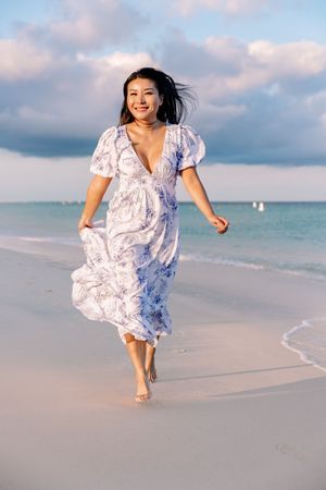 Woman in flowing blue floral dress running joyfully along pristine beach at sunset.
