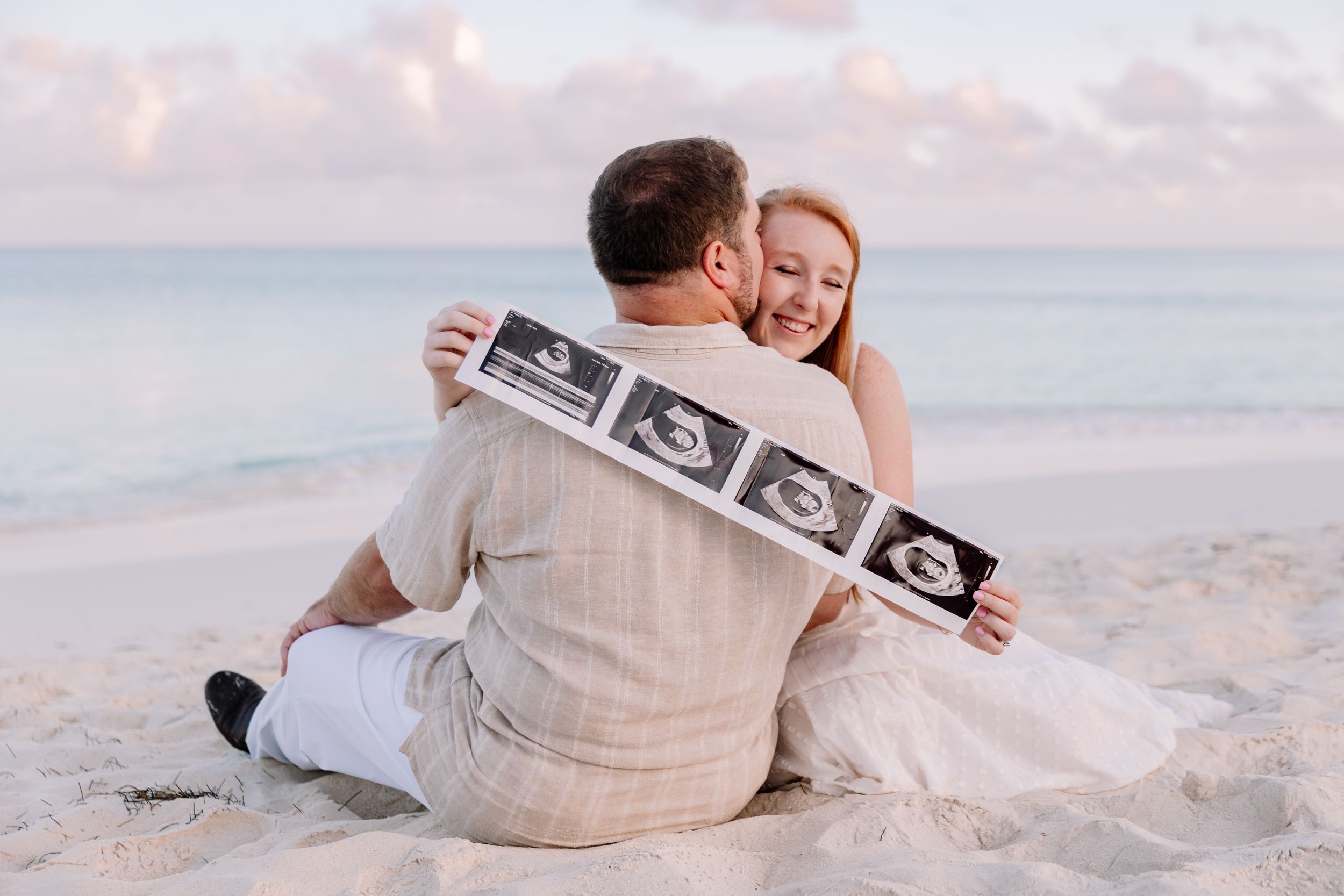 Couple on beach holding ultrasound photos while embracing.