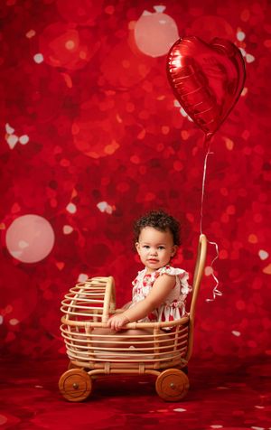 Small child sits in wooden toy wagon holding red heart balloon against vibrant red Valentine's Day backdrop.