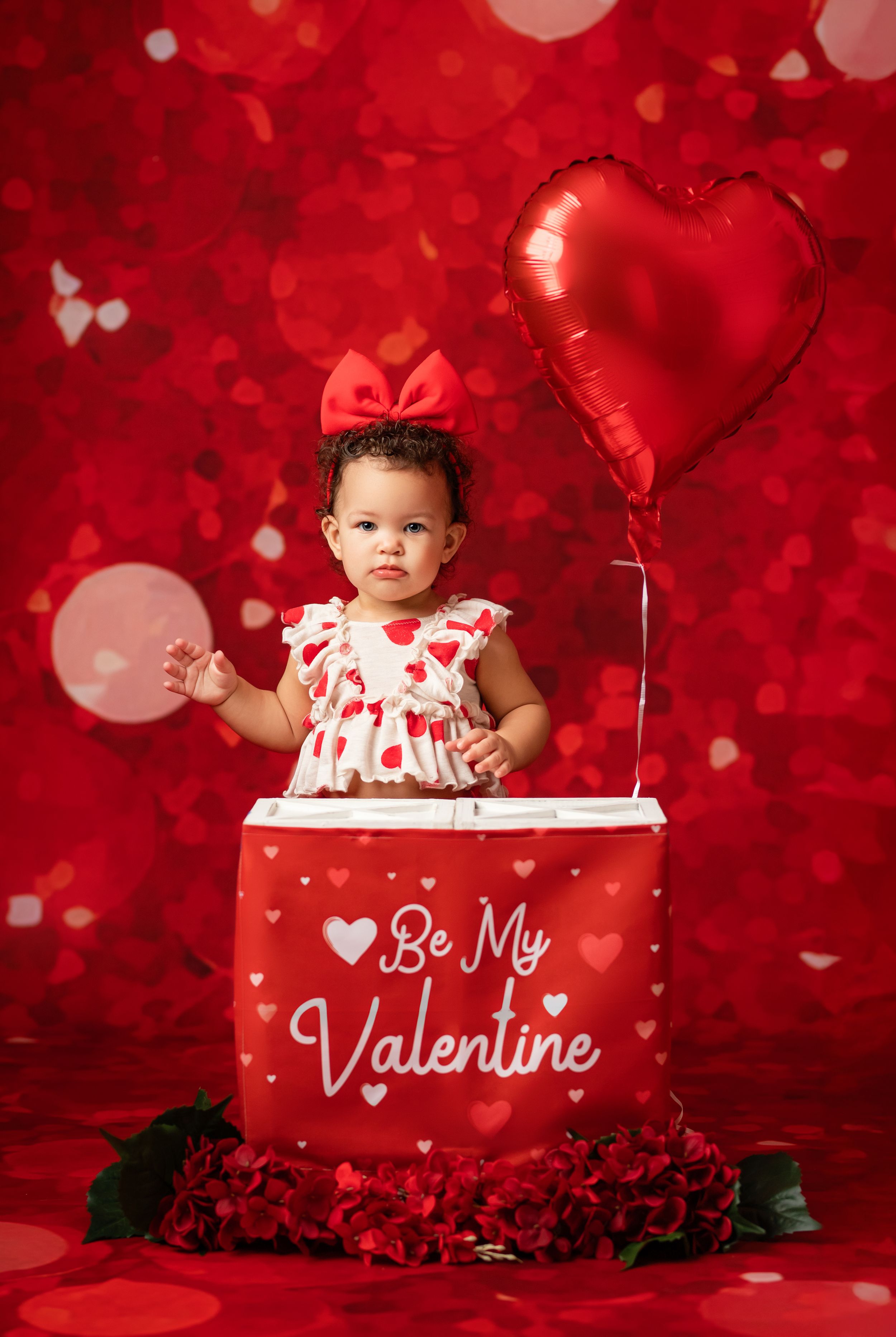 Baby in red and white Valentine's Day outfit poses in decorated red studio setting with heart decorations.