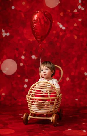 A child sits in a wicker basket with a red balloon against a festive red background with bokeh hearts and lights.