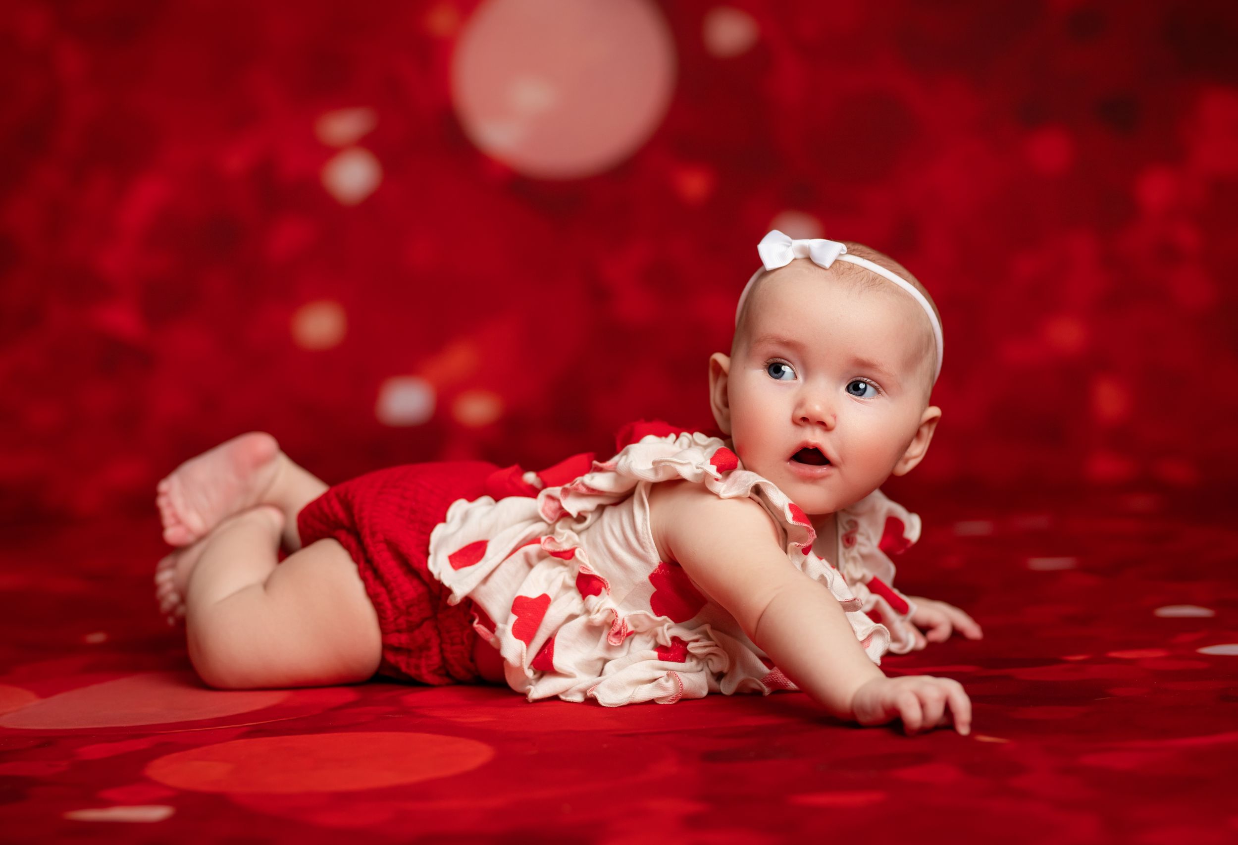 Baby in red and white floral outfit lies on plush red fabric background in creative photo series.