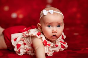 A baby in a white dress with red accents lies on a plush red background for a holiday portrait session.
