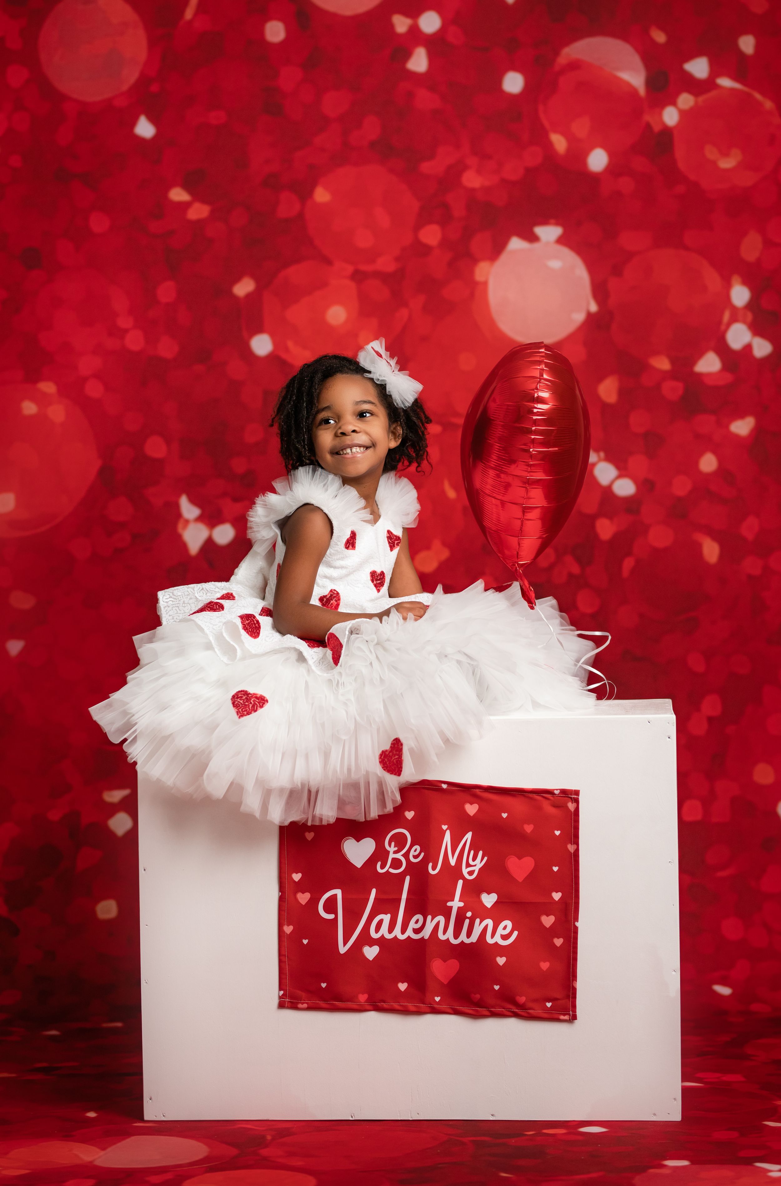 A Valentine's Day themed photo with a white tutu dress, red heart decorations, and a red balloon against a red backdrop.