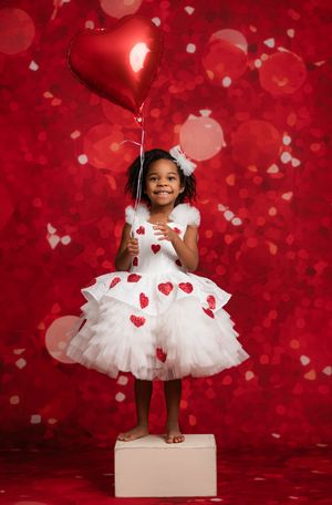 A child in a white tulle dress with red hearts holds a heart-shaped balloon against a red bokeh background.