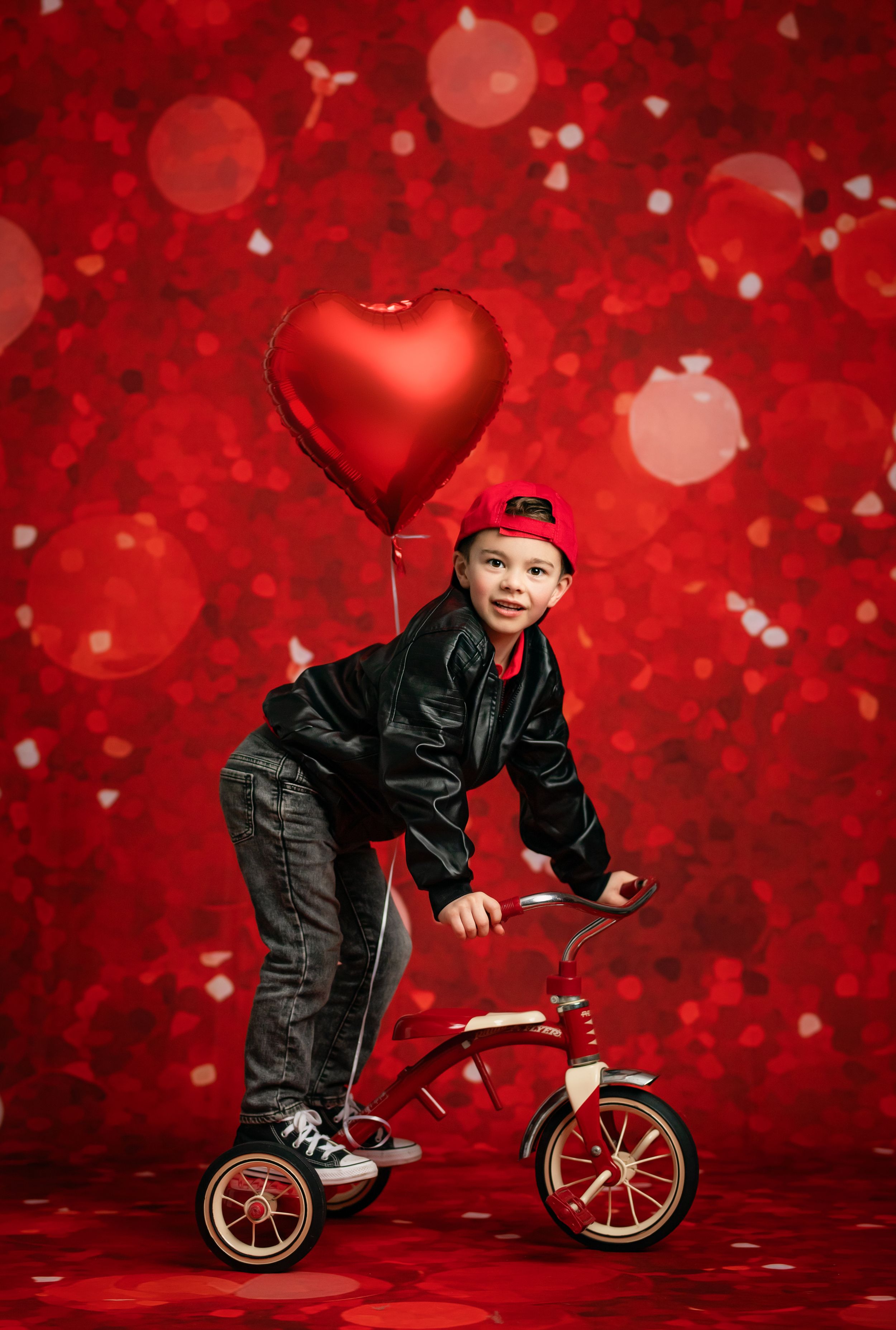 Child in black outfit rides red tricycle against vibrant red background with heart balloon and bokeh effects.