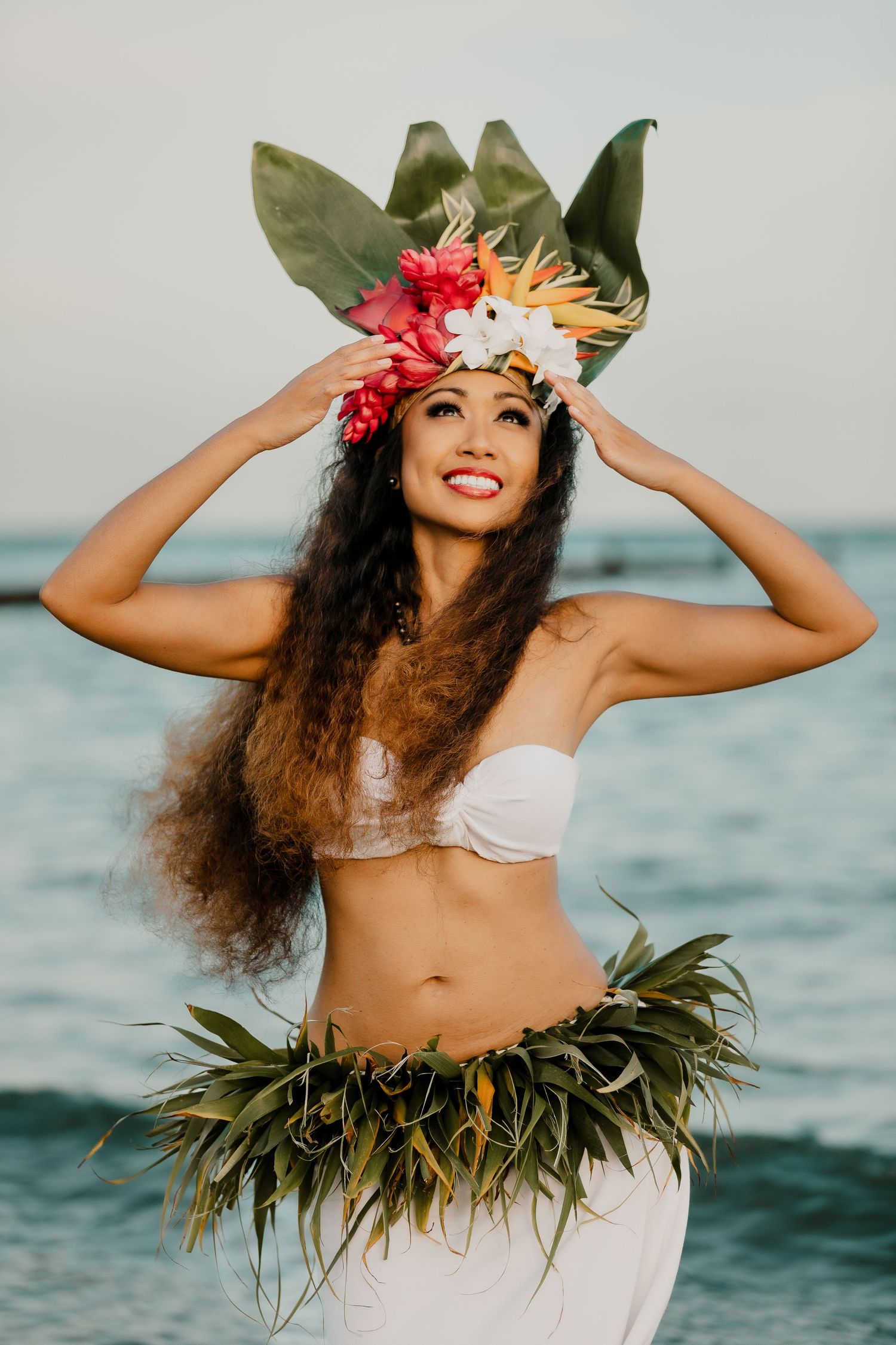 A dancer in traditional Hawaiian hula attire with tropical flowers and leaves poses gracefully by the ocean at sunset.