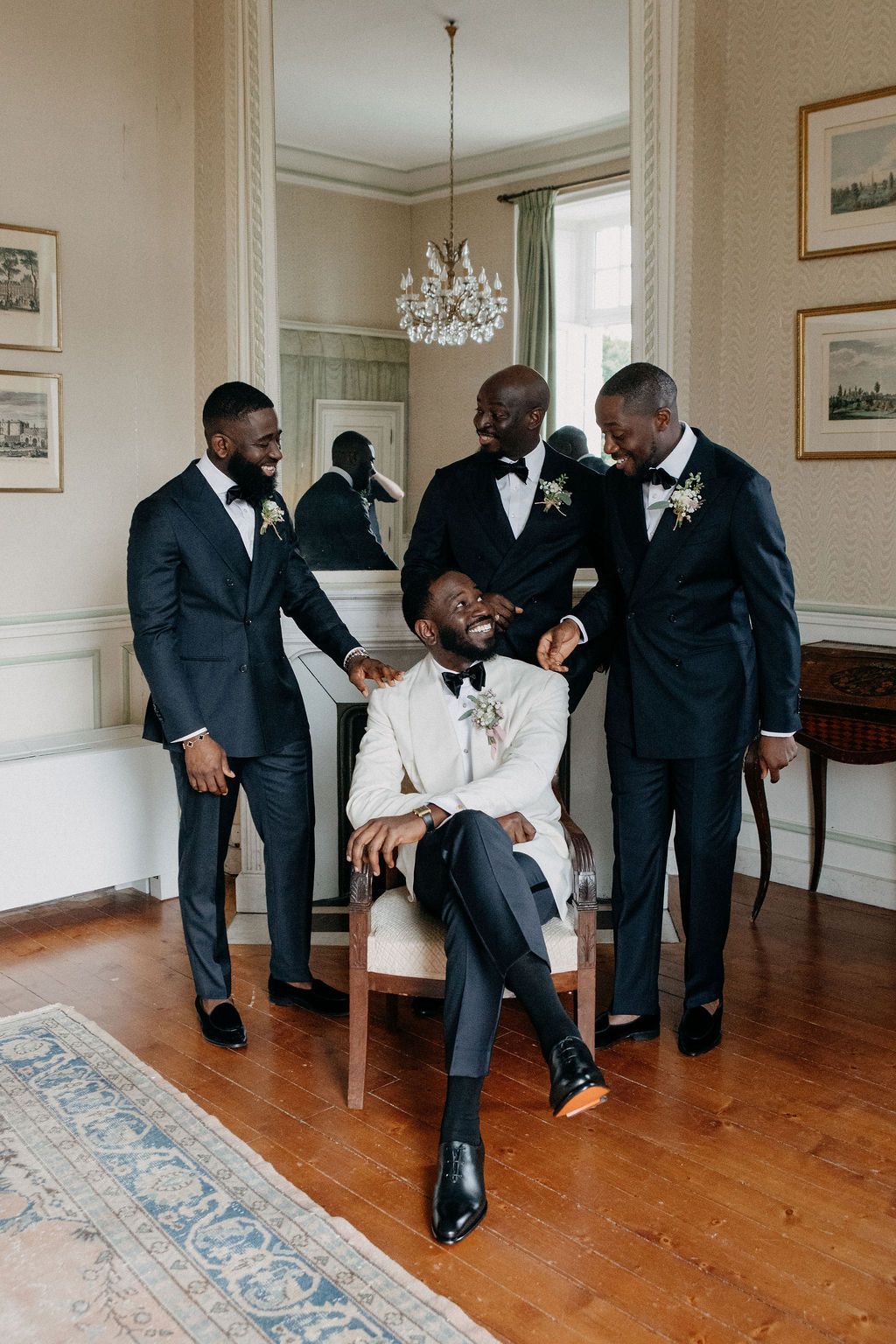 A laughing groom in a white tuxedo sits surrounded by three groomsmen in dark suits.