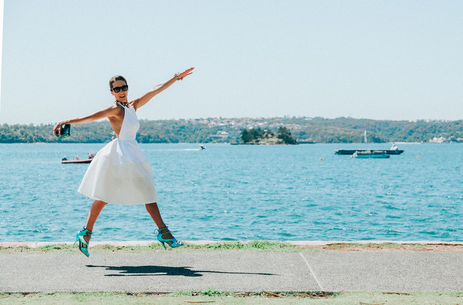 Person in white outfit striking graceful poses by waterfront with boats in background.