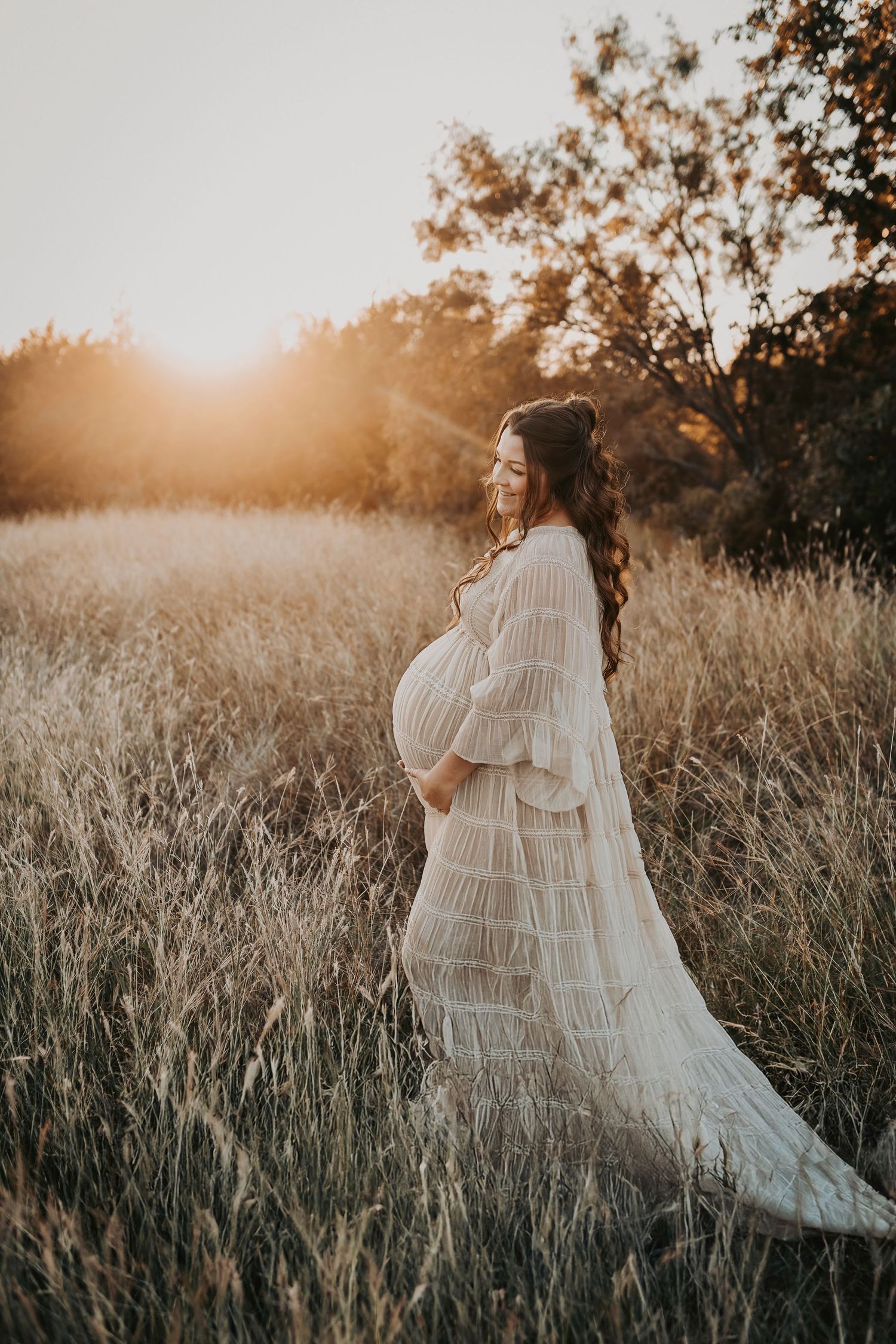 a golden glow falls over this pregnant mother as she holds her belly in a field of tall grass at sunset in dallas