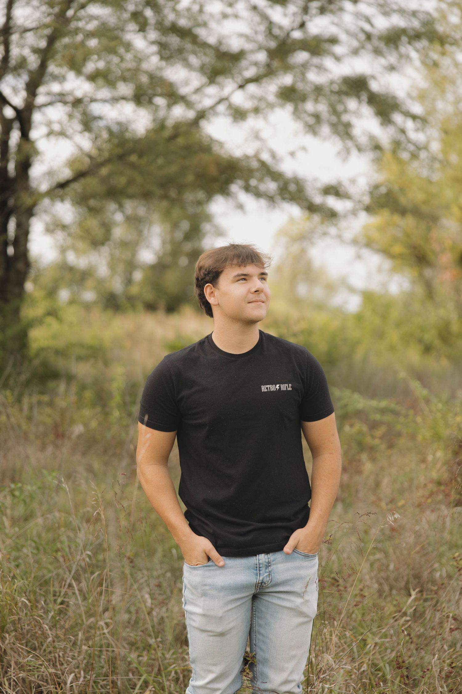 A person in a black t-shirt and light jeans stands in a field of tall grass during autumn.