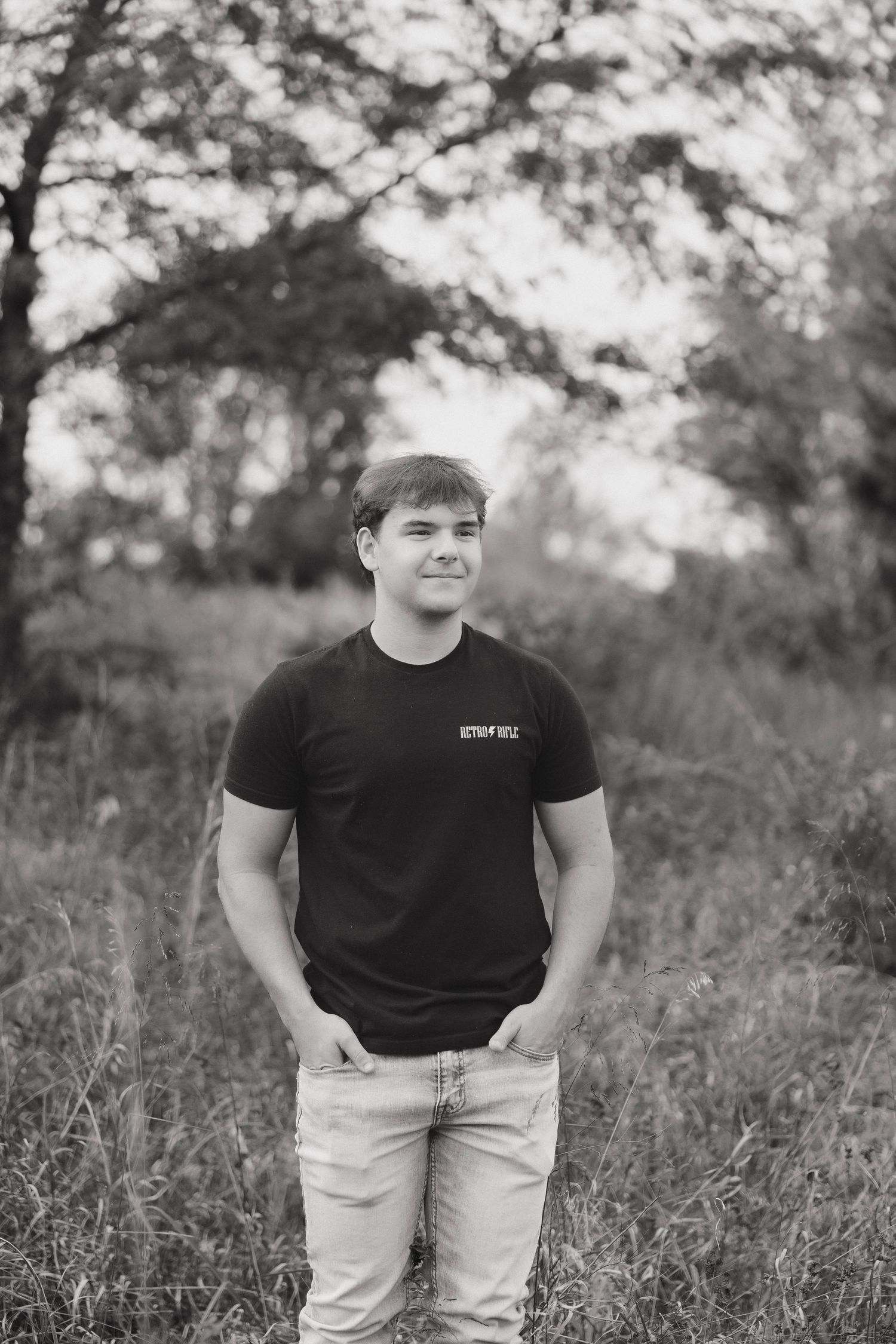 Black and white portrait of person in dark shirt standing in tall grass with trees in background.