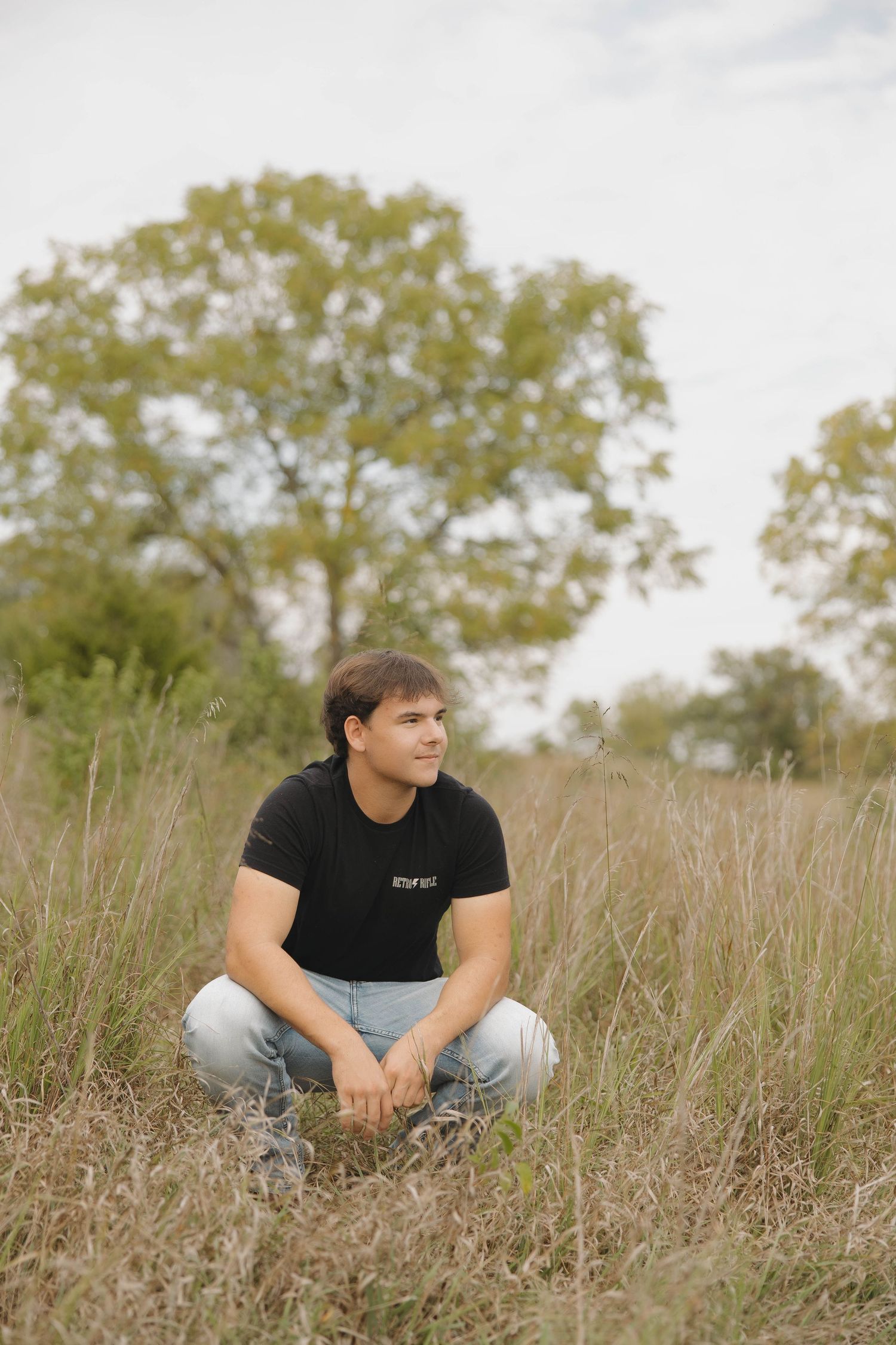 A person in a black t-shirt sits thoughtfully in a field of tall dried grass on a sunny autumn day.