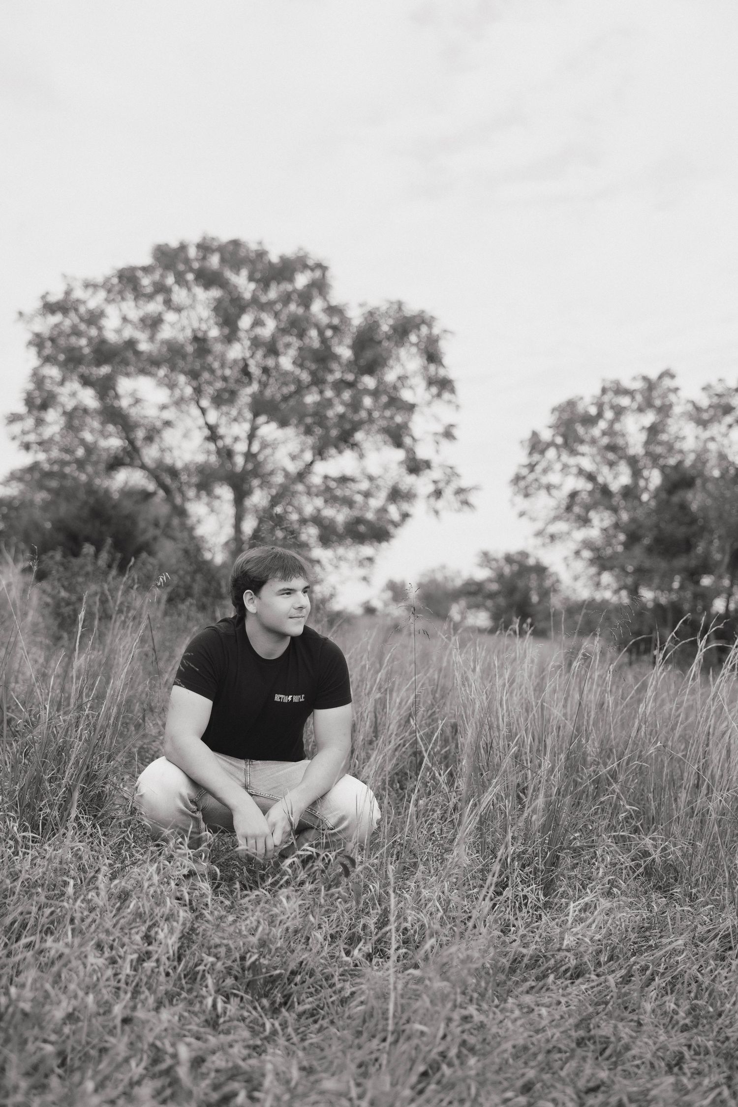 A person sits in a field of tall grass on a cloudy day in black and white.