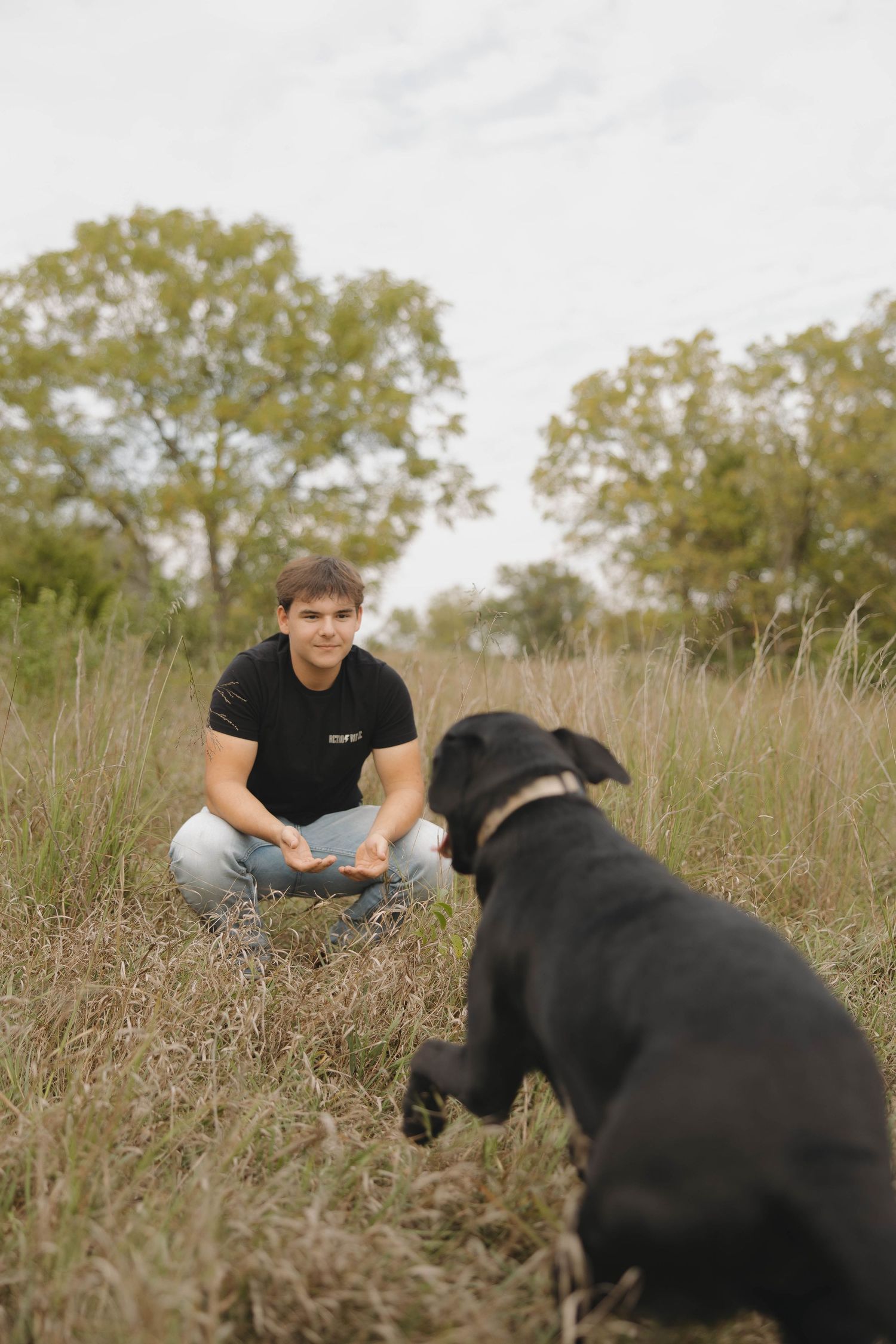 A person in a black shirt sits cross-legged in a grassy field with a black dog nearby.