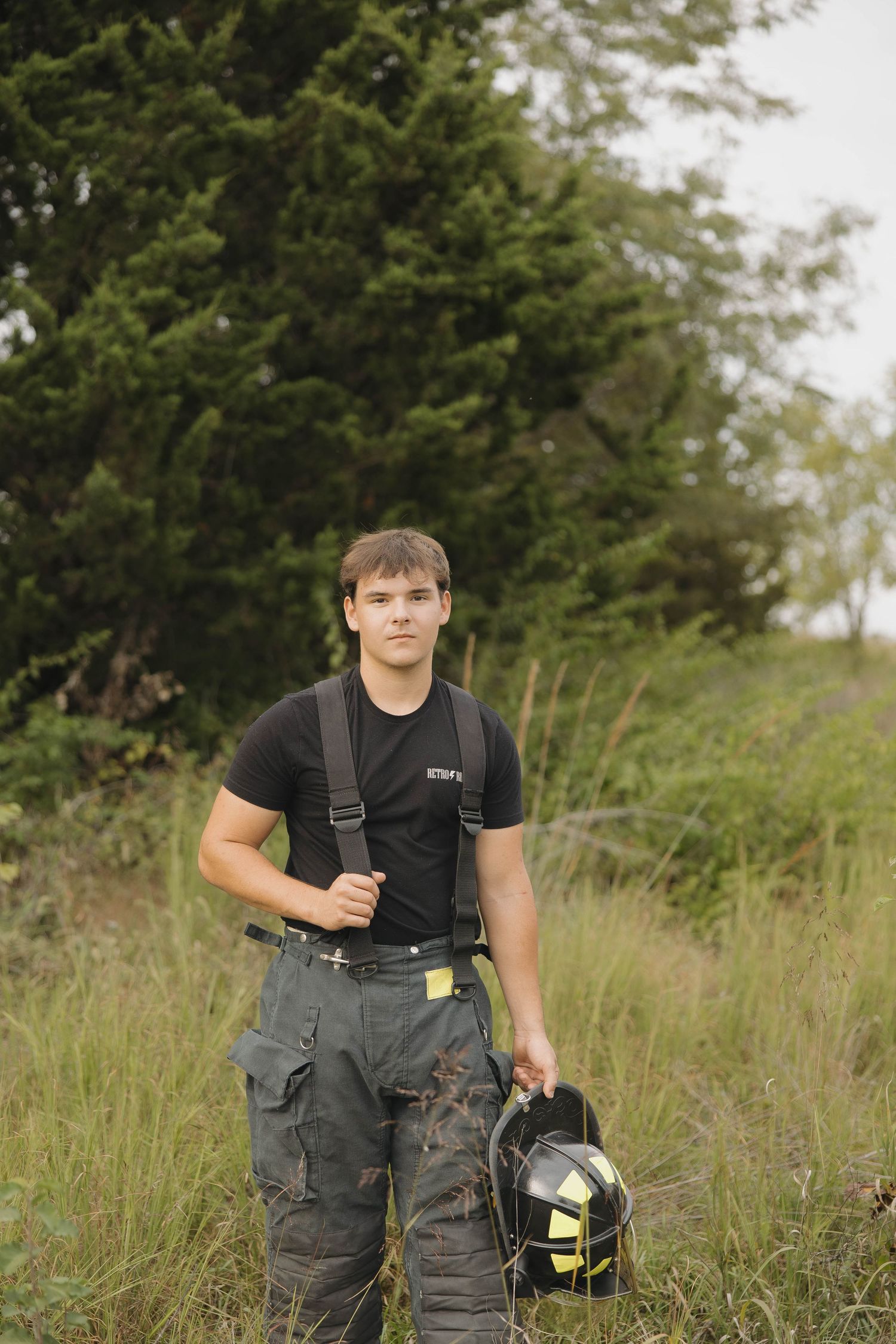 A firefighter in grey overalls and black shirt stands in tall grass holding protective gear.