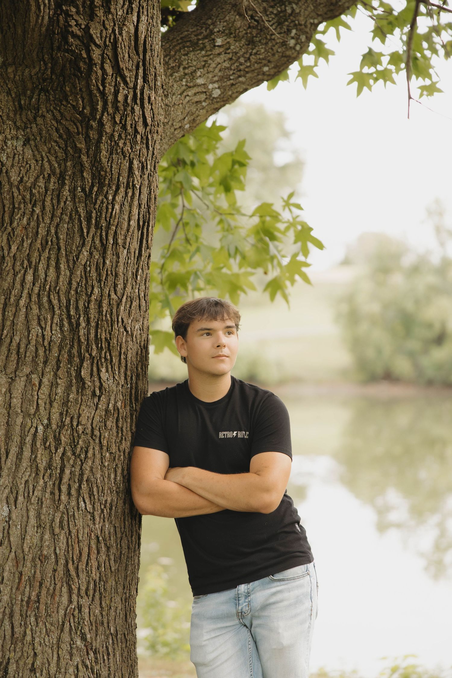 Person in black t-shirt and jeans stands with arms crossed by large tree trunk near peaceful lake.
